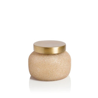 A beige candle in a round jar with a gold lid, set against a white background.
