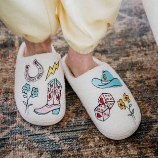 White slippers with colorful designs worn by a person in light-colored pants on a textured floor.
