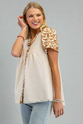 Woman wearing a beige top with floral sleeves and light blue jeans on a gray background
