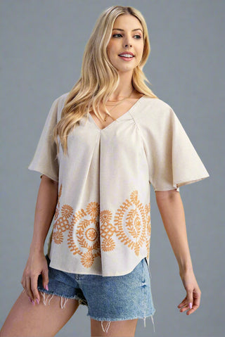 Woman wearing a beige top with gold embroidery and denim shorts on a gray background