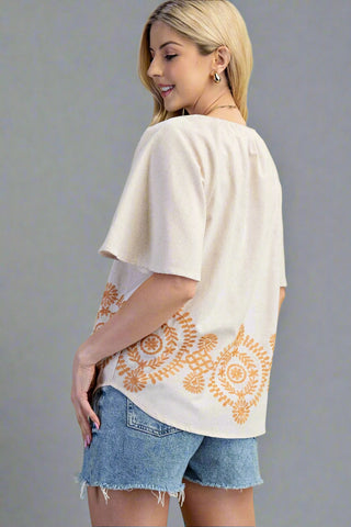 Woman wearing a beige top with orange floral patterns and denim shorts on a gray background