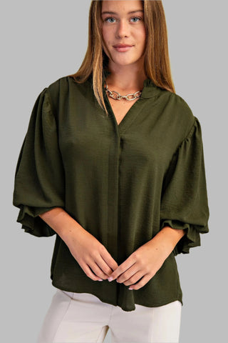 Woman wearing an olive green blouse with white pants against a light green background